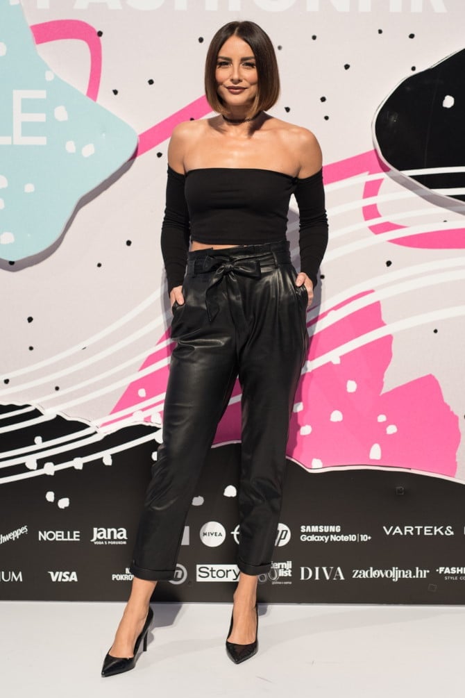 Woman in black off-shoulder top and leather pants posing at event with colorful pink and black backdrop