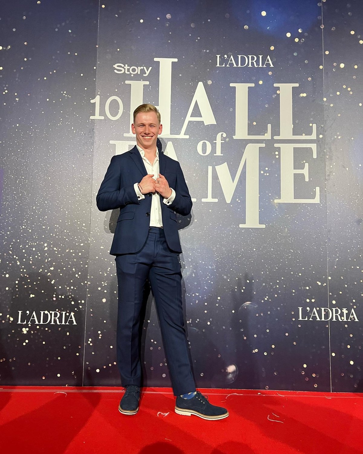 Man in dark blue suit smiling on red carpet at Hall of Me premiere event with sparkly backdrop