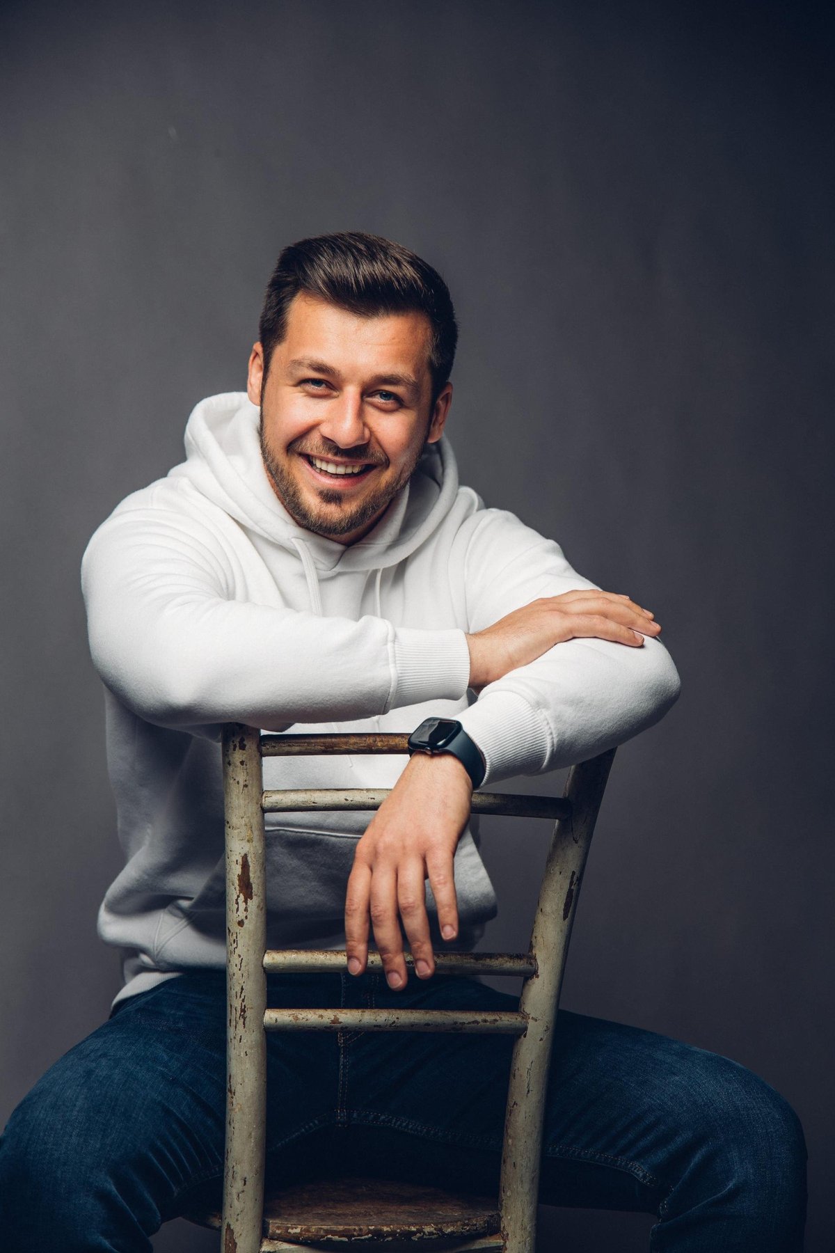 Alen Đuras, hrvatski pjevač i natjecatelj Plesa sa zvijezdama 2026., nasmijan u bijeloj majici s kapuljačom sjedi na drvenoj stolici – Plesna škola Banderillas Zagreb
