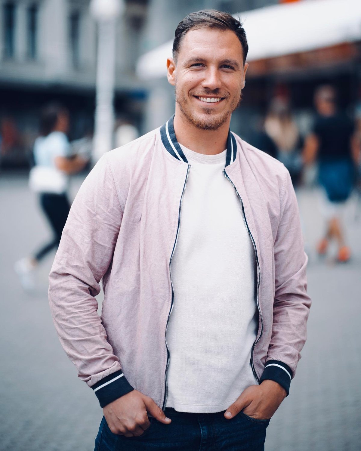 Smiling man wearing pink bomber jacket and white t-shirt standing outdoors in urban setting