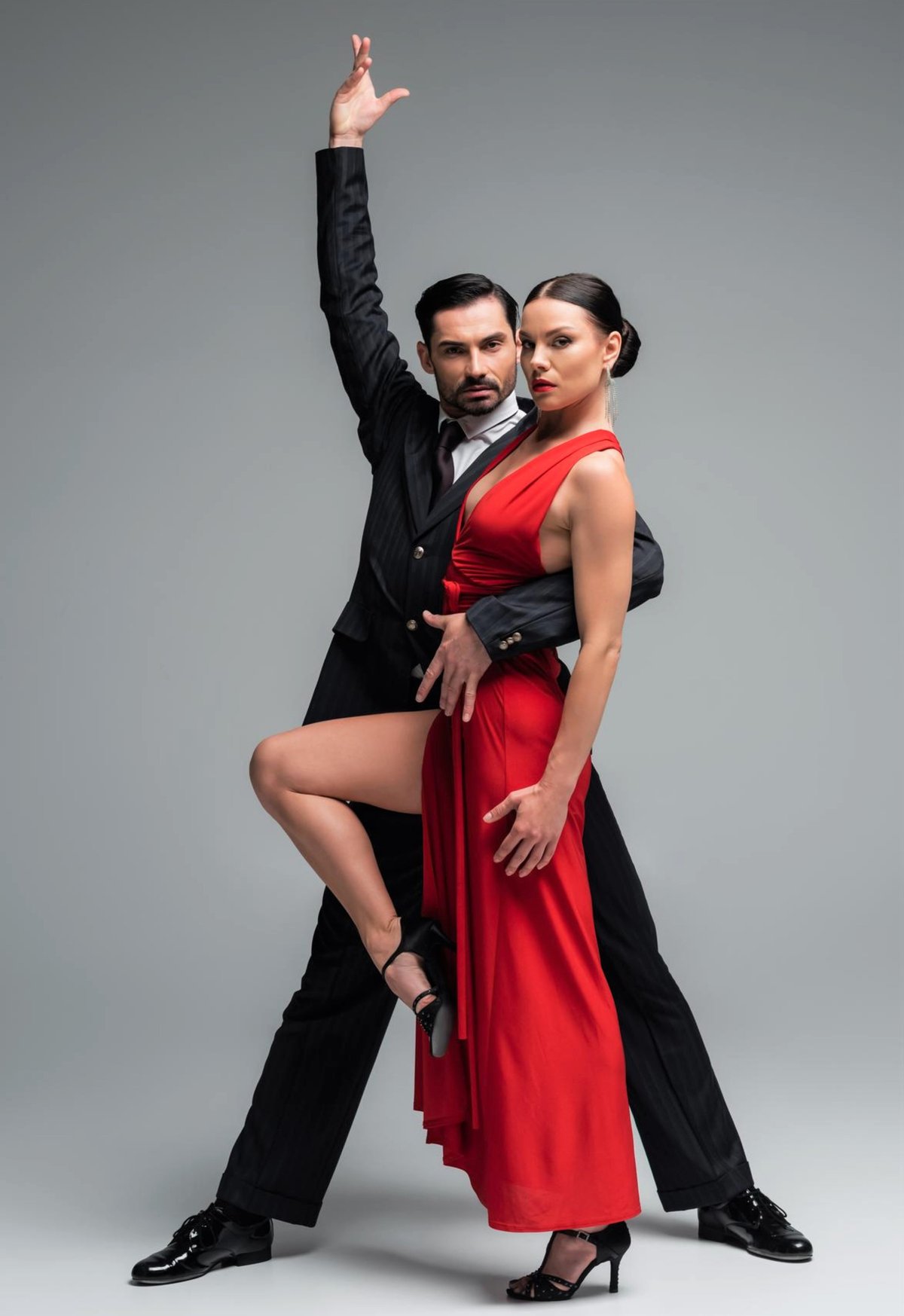 Man in black suit and woman in red dress performing a dramatic dance pose together against a gray background