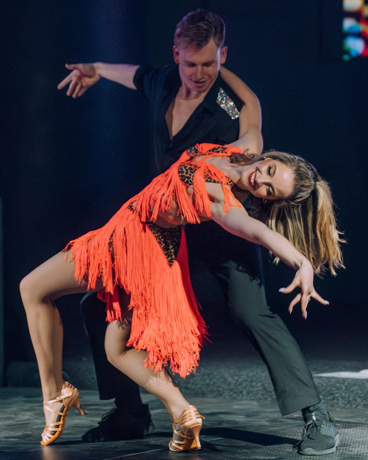 Male and female dancers performing Latin dance in elegant costumes on stage with dark blue background