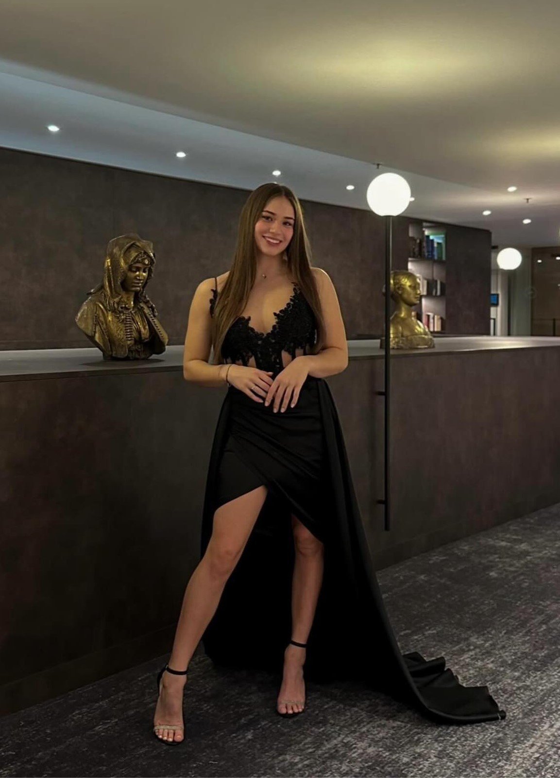 Woman in black velvet dress posing in modern hotel lobby with bronze bust sculpture and ambient lighting