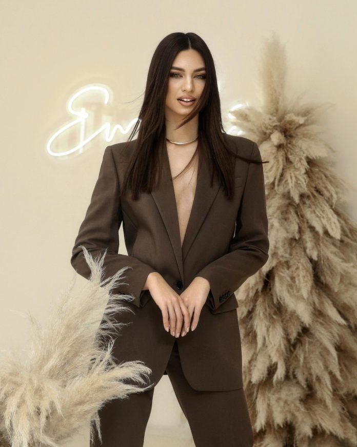 Woman with long dark hair wearing a brown blazer, posing between pampas grass with a neon sign in the background
