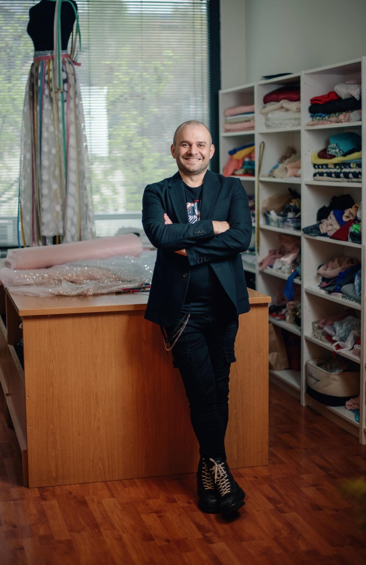 Man leaning against wooden counter in studio with arms crossed, wearing black suit. Shelves with clothing and sneakers visible behind him.